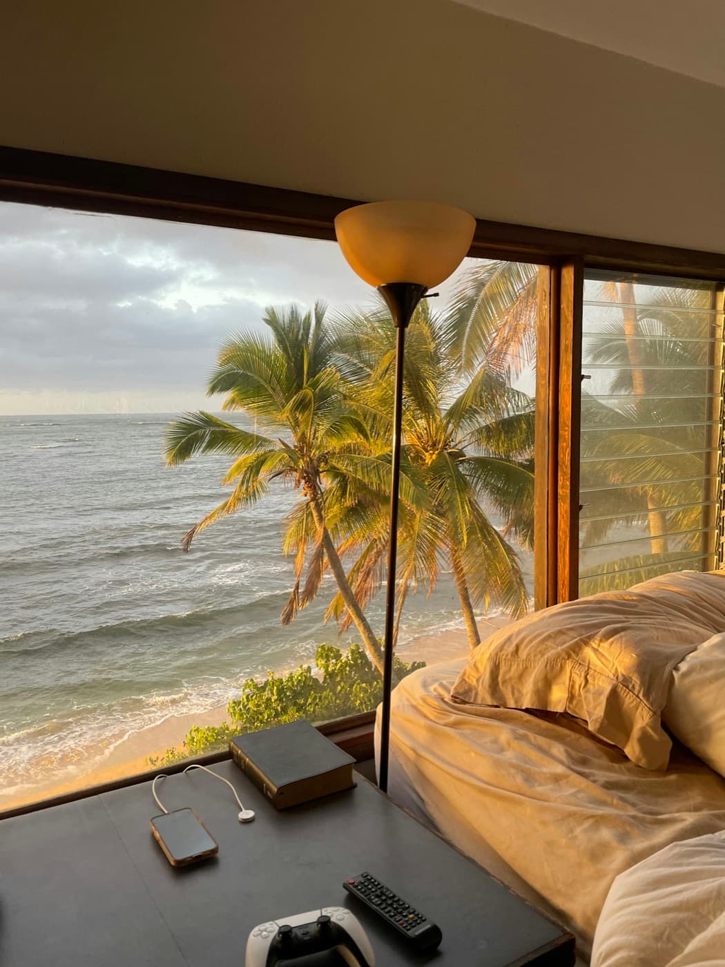 Quarto com a janela aberta para o mar e coqueiros ao pôr do sol.
